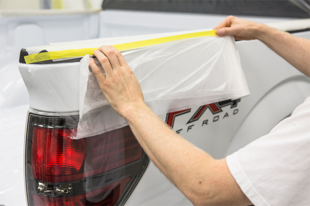 PlazMask maskign film being applied to the back right of a pick-up truck. The taping process is just beginning as a man is applying the yellow tape to the truck cab, directly above the back right taillight.