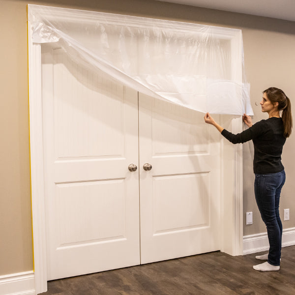 A woman pulling the transparent pre-taped masking film from the top of a white door frame down towards the bottom to protect the white doors from paint.