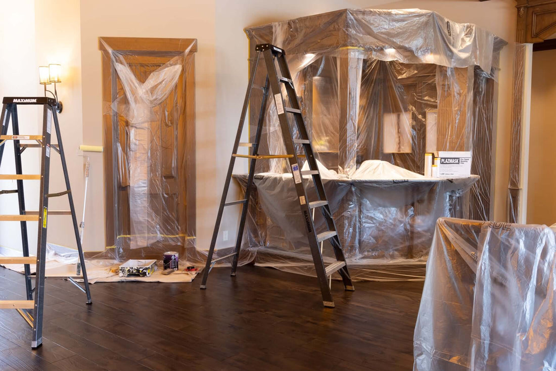 two ladders; one medium height and one tall, stand in an open living space with brown hardwood floors and white walls. There is a brown bar in the back right, a brown wooden door to the left of the bar, and a chair in the right foreground covered in masking film to protect from paint.