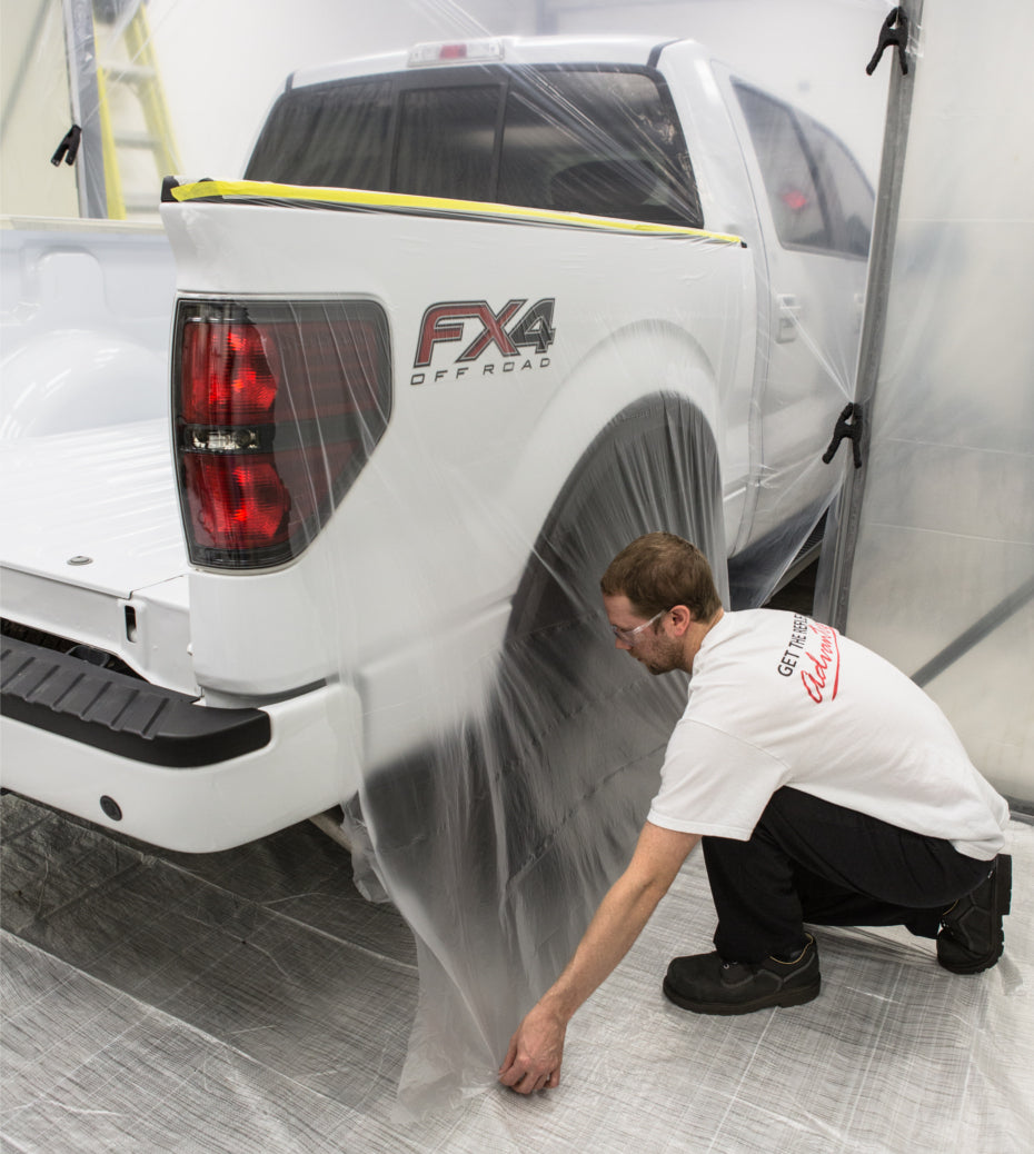 A man in a white shirt and black pantsis fastening the PlazMask masking film to the ground after covering the back right of his truck with the film.